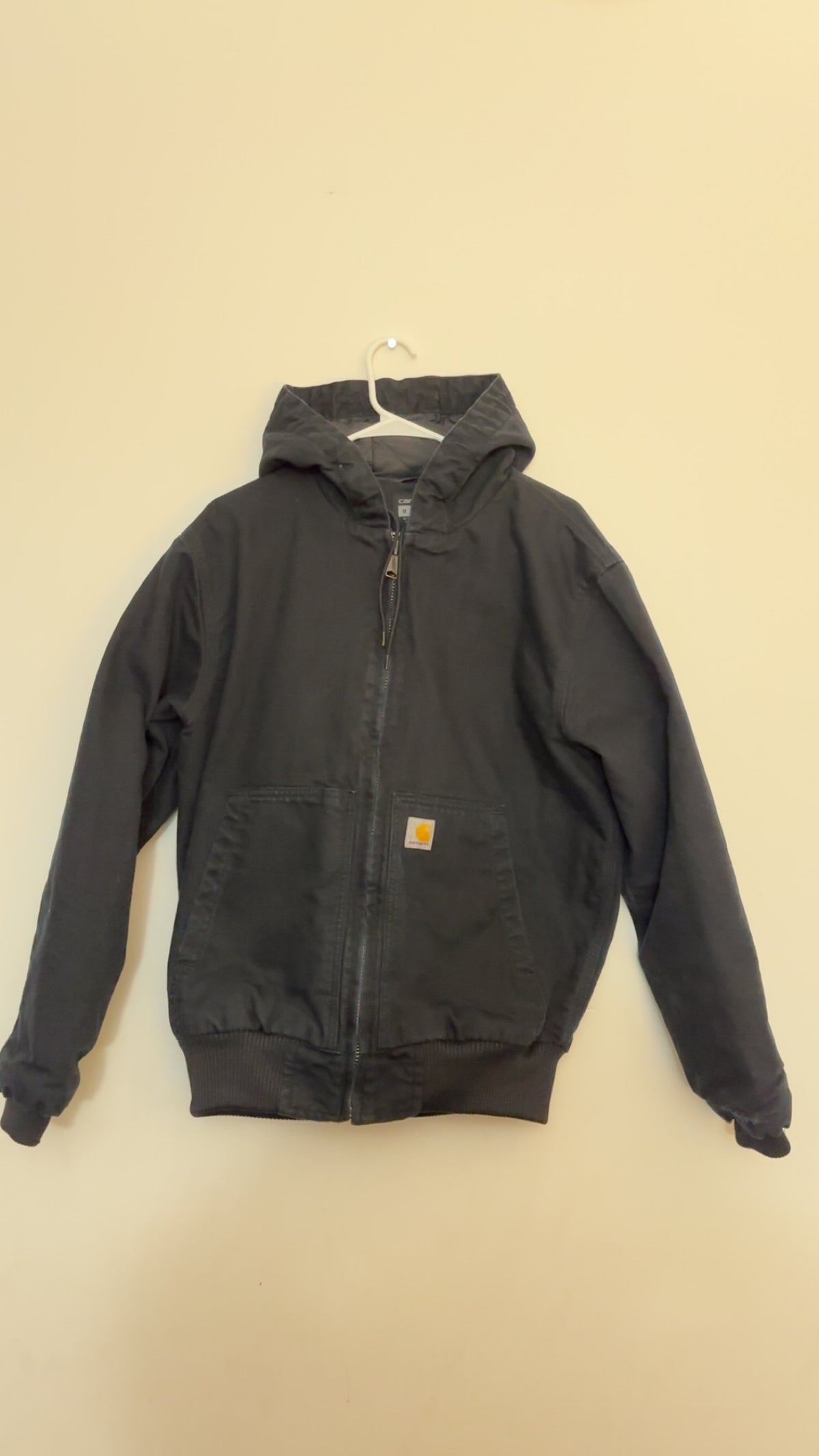 A black Carhartt hooded jacket with a front zipper and a small orange Carhartt logo patch on the chest, 